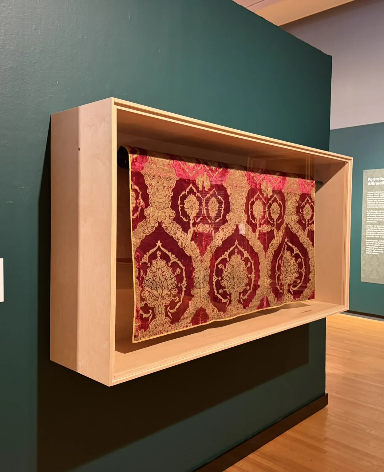 A wall-mounted maple vitrine holding a red and gold Renaissance textile