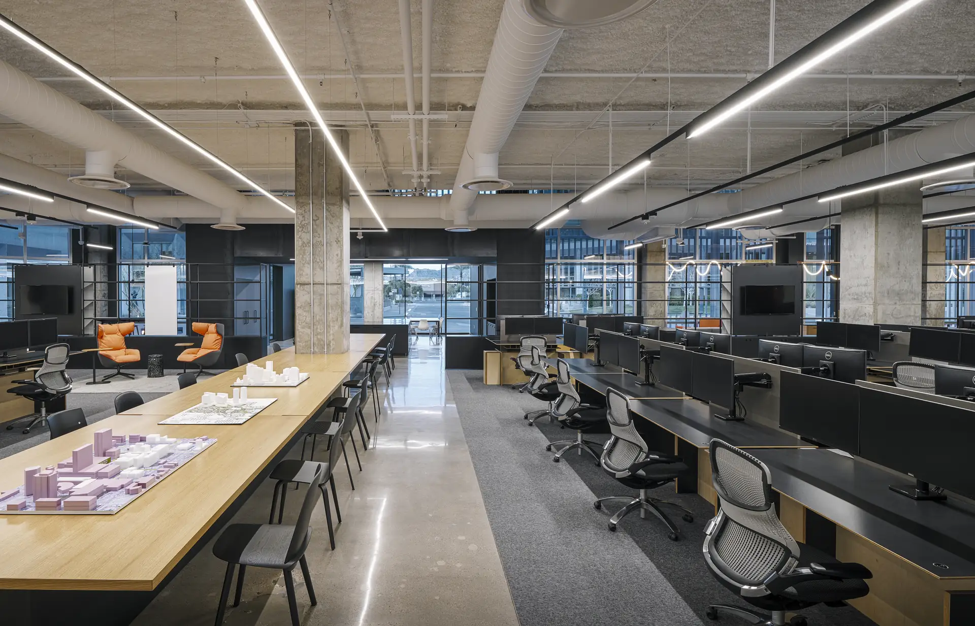 An open office floor with benching workstations and a long shared table under linear lighting
